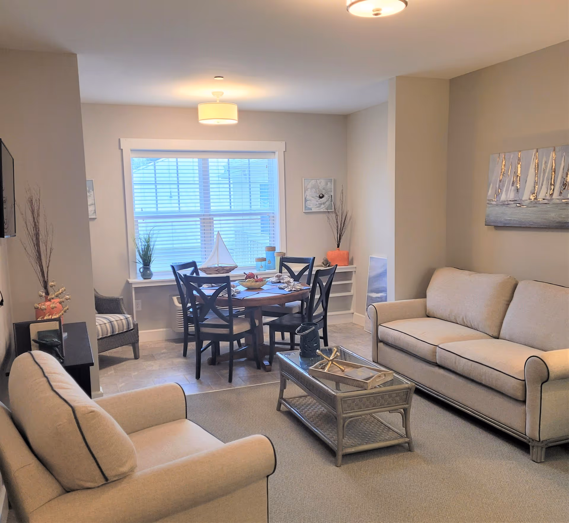 A cozy living room and dining area with beige walls and carpet. The living room has a beige sofa and armchair with dark piping, a glass-top coffee table with decorative items, and a painting of sailboats on the wall. The dining area features a round wooden table with four chairs, a window with blinds, and nautical-themed decorations including a model sailboat and coral pieces.