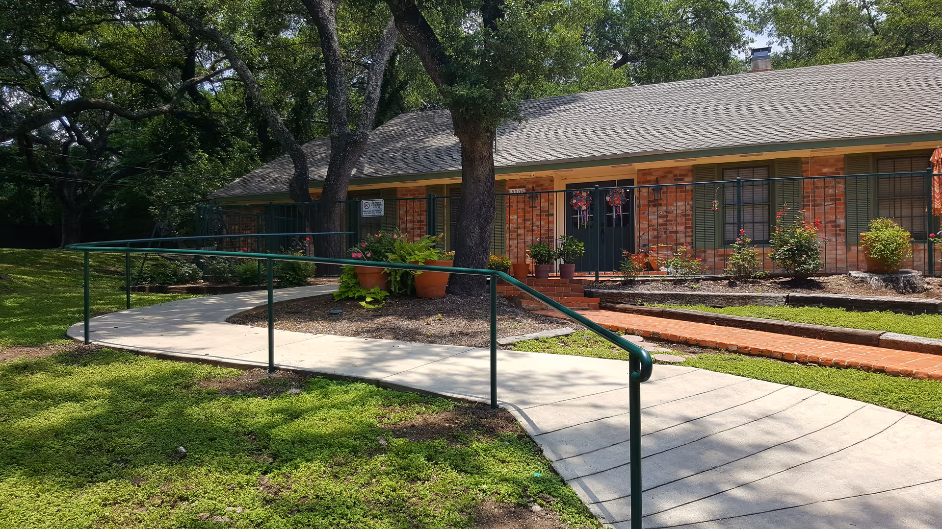 A single-story brick building with green shutters and a gray shingled roof, surrounded by greenery and trees. A concrete wheelchair-accessible ramp with green handrails leads up to the entrance, which has double doors decorated with wreaths. There are potted plants and flower beds near the entrance.