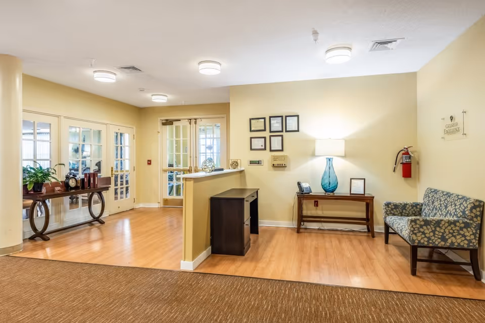 Reception area with a small desk and a table with a blue glass lamp, a phone, and framed certificates on the wall. There is a patterned armchair and a fire extinguisher mounted on the wall. Double glass doors and a wooden table with decorative items and a plant are visible in the background.