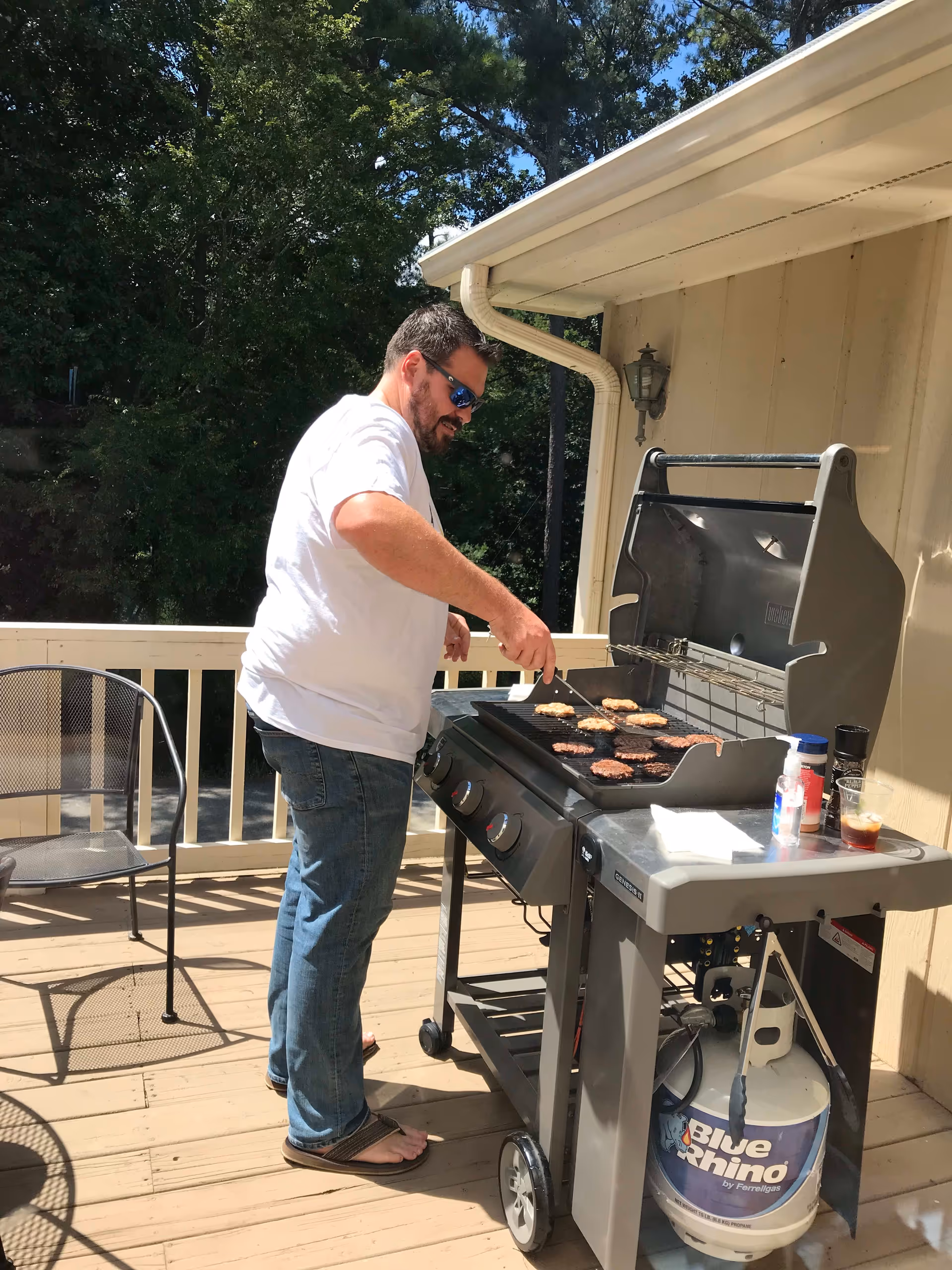 Person grilling burgers on a gas grill on a sunny wooden deck attached to a house.