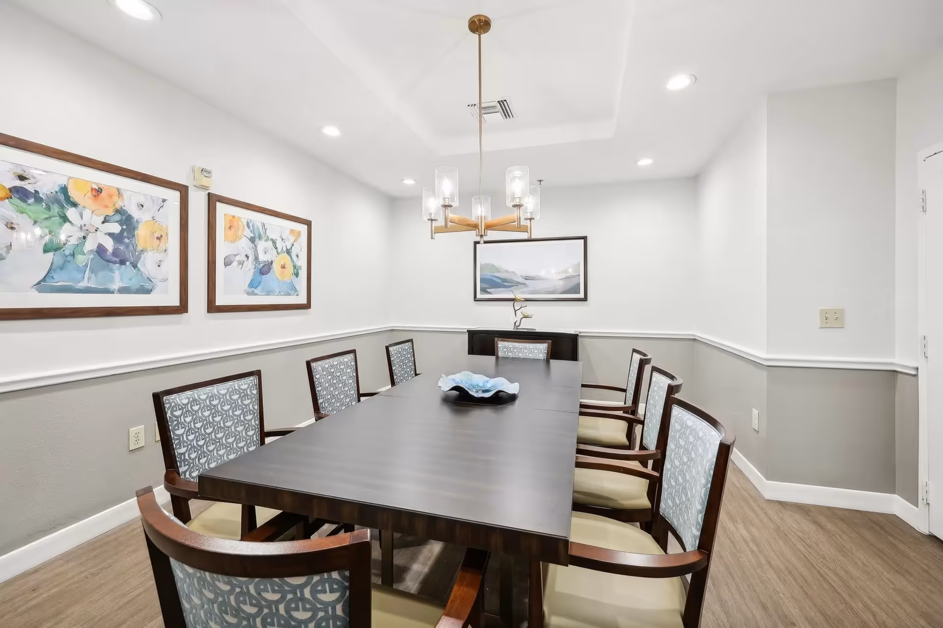 A well-lit dining room with a long dark wooden table surrounded by eight cushioned chairs with patterned backs. The walls are painted white and gray with three framed floral and landscape paintings hanging. A modern chandelier with glass shades hangs above the table, and the floor is wood.