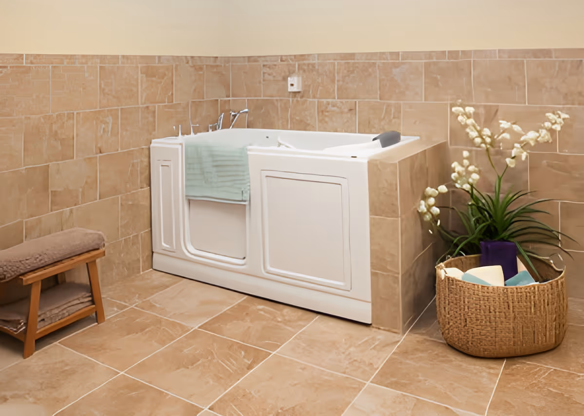 A walk-in bathtub with a door, surrounded by beige tiled walls and floor. A light green towel hangs over the edge of the tub. To the left, there is a small wooden bench with folded towels on the lower shelf and a cushioned seat. To the right, a woven basket contains bath items and a plant with white flowers.