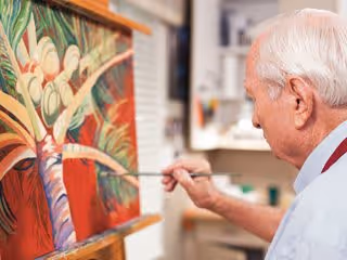An elderly man painting a colorful tropical scene on a canvas in an art studio or creative space.