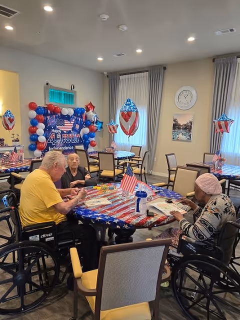 Three elderly individuals sitting around a table decorated with a red, white, and blue American flag-themed tablecloth and small American flags. The room is decorated with patriotic balloons and a banner that reads 'Happy 4th of July Independence Day'. The setting appears to be a communal dining or activity area in a senior living facility.