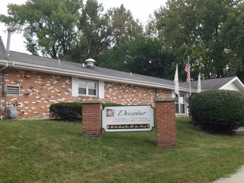 Brick facility exterior with a sign reading Decatur Rehabilitation Health Care Center and flags.