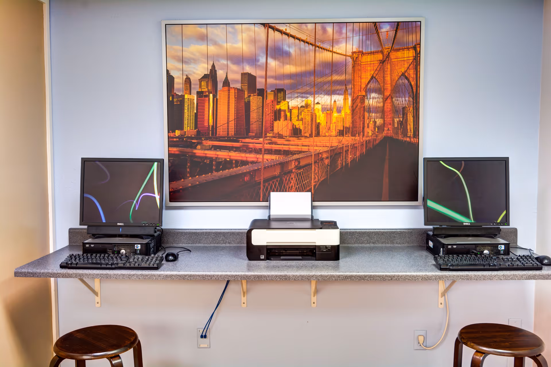 A small computer workstation area with two desktop computers and a printer on a long counter. Two wooden stools are placed in front of the counter. Above the counter is a large framed picture of a city skyline and a bridge at sunset.