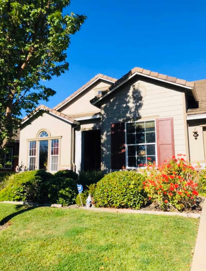 Exterior view of a single-story house with beige siding, brown roof tiles, and red shutters. The front yard features green grass, bushes, and red flowers under a clear blue sky.