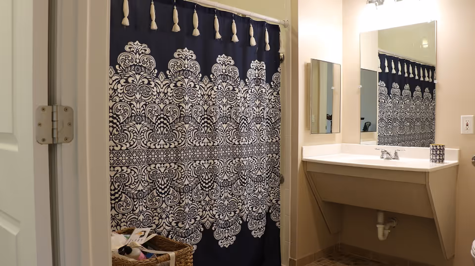 Bathroom with a decorative dark blue and white patterned shower curtain, a white sink with a mirror above it, a small wall-mounted cabinet, and a basket with toiletries on the floor.