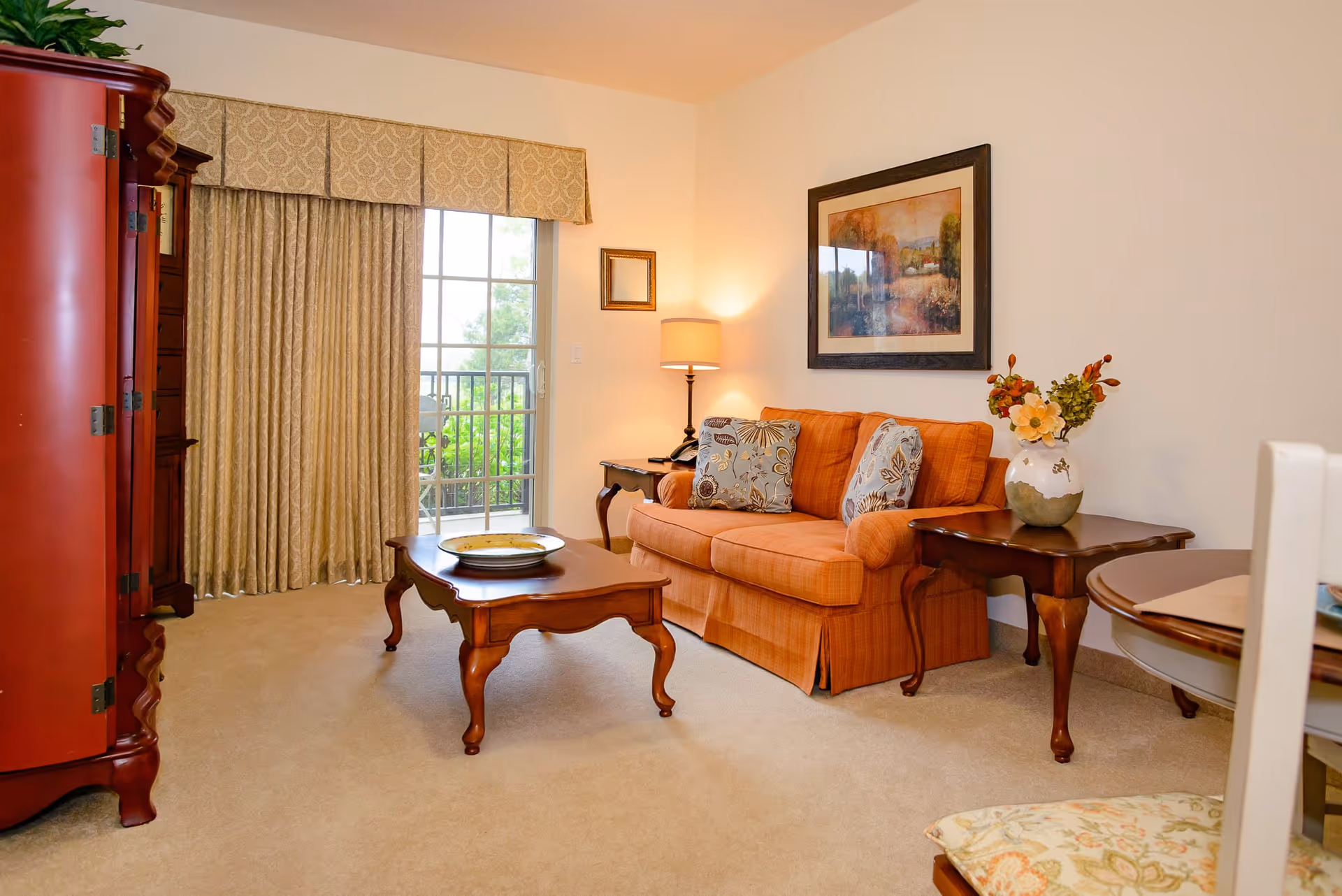 A cozy living room with an orange sofa adorned with patterned cushions, a wooden coffee table with a decorative plate, a side table with a vase of flowers, a floor lamp, and a framed painting on the wall. There is a large window with beige curtains leading to a balcony with greenery outside. A red wooden cabinet is partially visible on the left side.