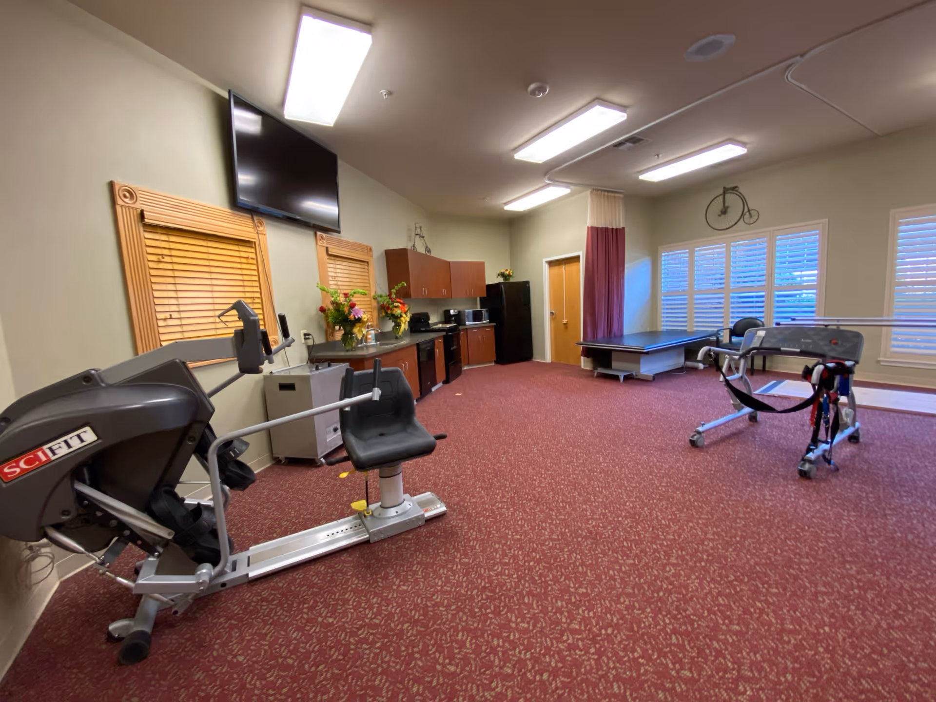 Well-lit interior therapy room with exercise equipment, a treatment table, kitchenette cabinets, and a wall-mounted TV.