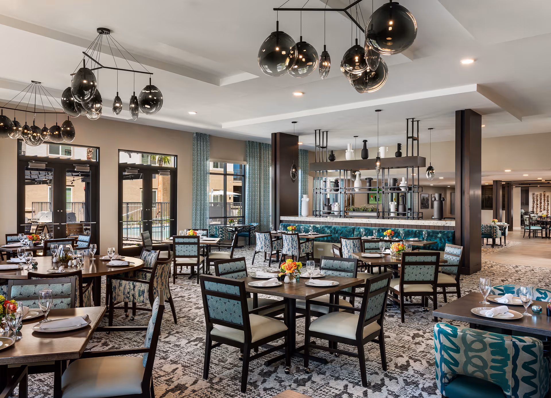 A spacious and modern dining room with multiple tables and chairs arranged neatly. The tables are set with plates, glasses, and napkins, and decorated with small flower arrangements. Large windows and glass doors allow natural light to enter, and contemporary pendant lights hang from the ceiling. The room features patterned carpeting and a mix of upholstered chairs and banquette seating along the walls.