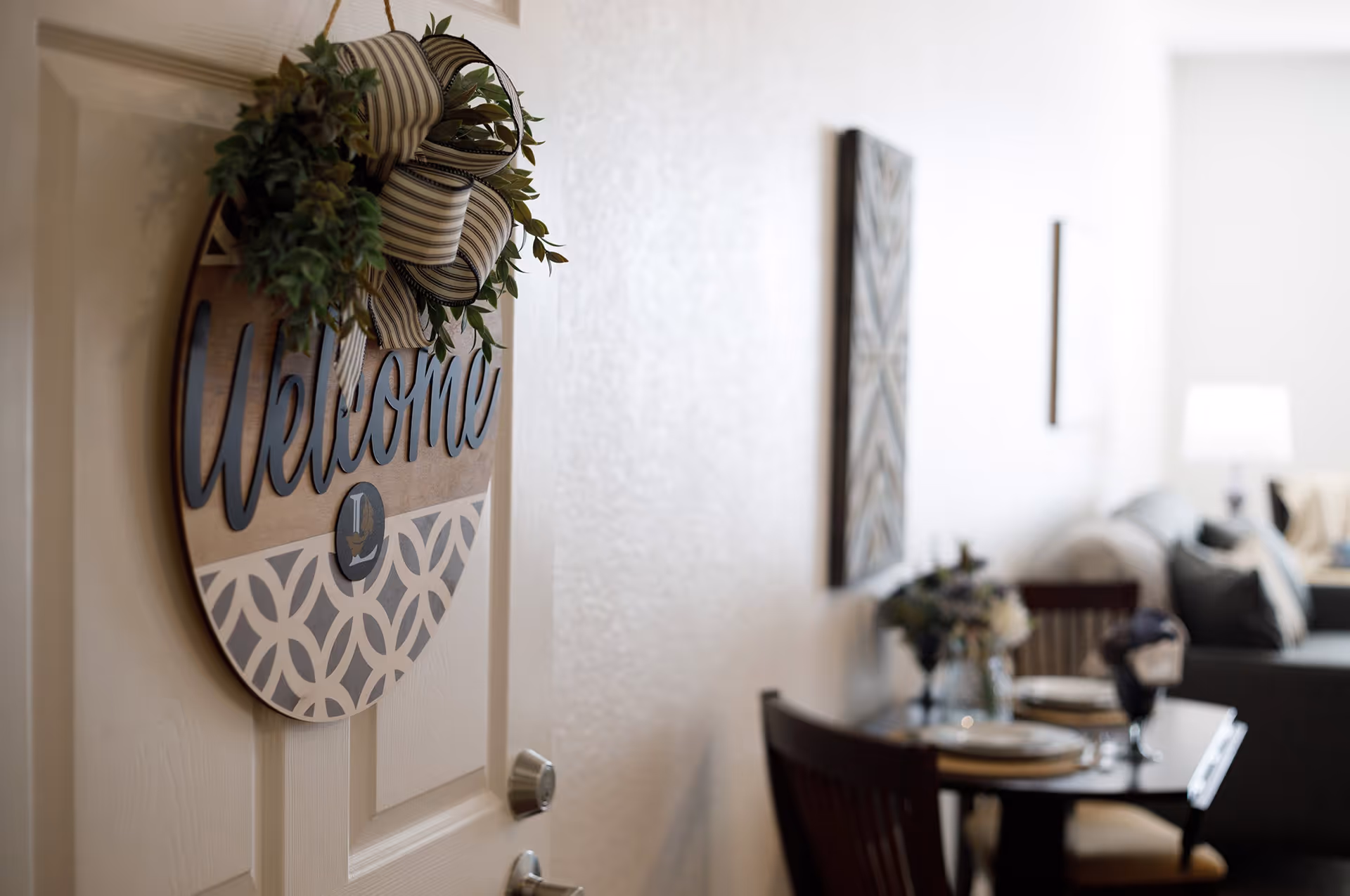 Close-up of a door with a decorative 'Welcome' sign featuring a striped bow and greenery. In the background, a dining table set with plates and glasses is visible, along with a living area that includes a couch and a lamp.