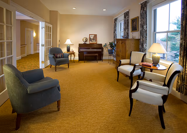A cozy living room area with beige carpet and cream-colored walls. The room features a dark wooden piano against the far wall, a small side table with a lamp and a globe, two blue upholstered armchairs, and two white cushioned chairs with dark wooden frames around a small round table with a lamp and books. Large windows with patterned curtains allow natural light to enter the room.