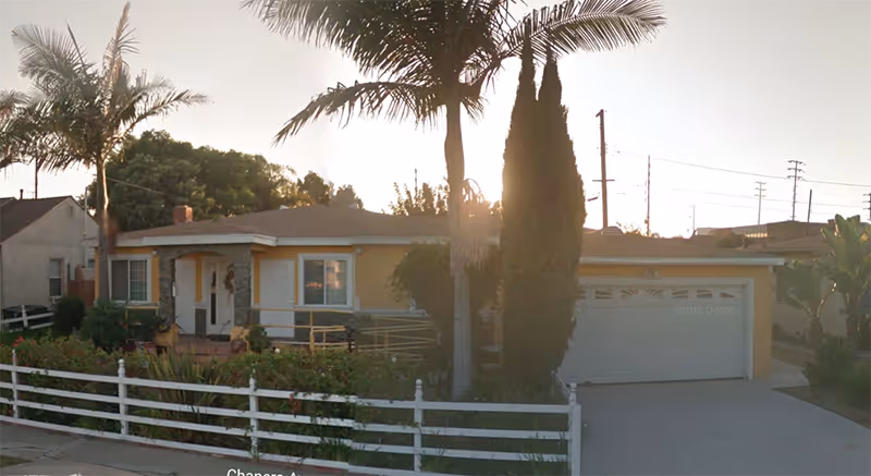 Front view of a single-story yellow villa with a white fence and palm trees.