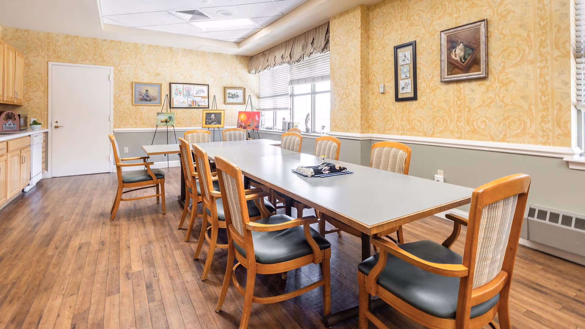 A well-lit dining room with a long rectangular table surrounded by wooden chairs with cushioned seats. The room has wooden flooring, yellow patterned wallpaper, and several framed artworks on the walls. There is a window with blinds letting in natural light and a counter with cabinets on one side.