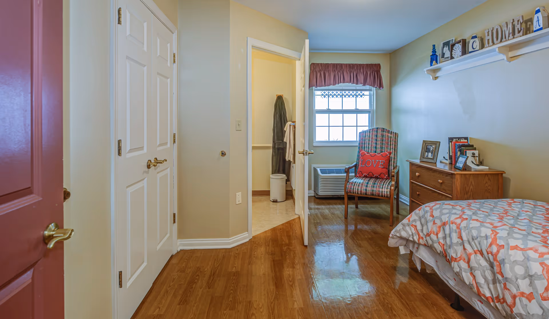 A cozy bedroom with a single bed covered in a patterned comforter, a wooden dresser with framed photos and books, a plaid armchair with a red pillow that says 'LOVE', a window with a red valance, and a door leading to a bathroom. The walls are painted light yellow and the floor is wooden.