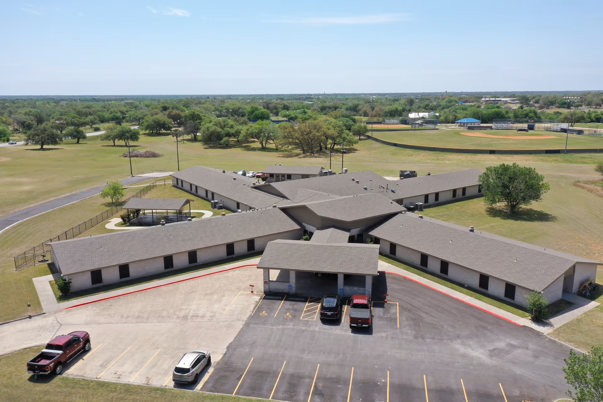 Aerial view of a single-story nursing and rehabilitation facility with interconnected wings, a central entrance, a parking lot, and surrounding green fields.