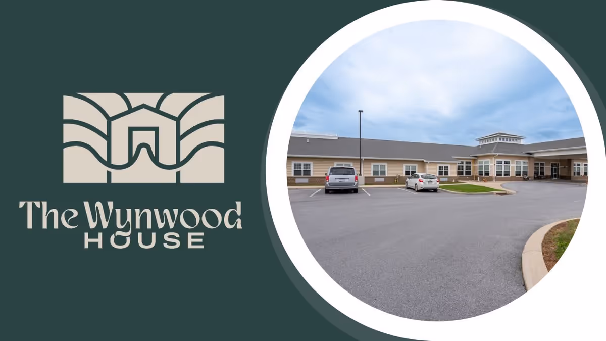 Exterior view of The Wynwood House personal care home building with a parking lot in front, two cars parked, and a cloudy sky overhead.