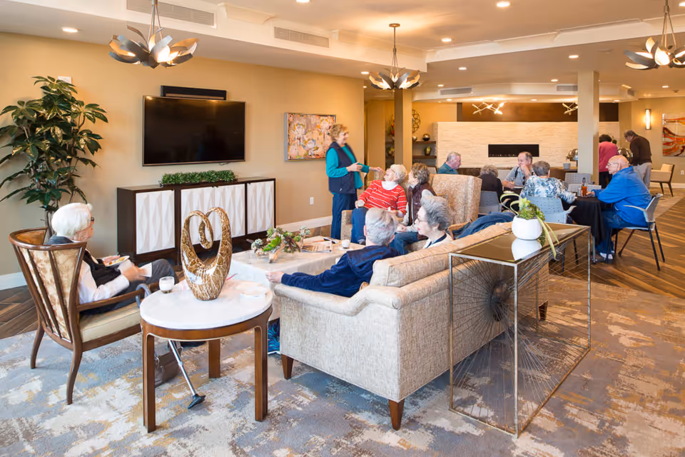 A group of elderly people socializing in a well-lit living room with modern decor. The room features a beige sofa, armchairs, a flat-screen TV mounted on the wall, decorative plants, and a coffee table with a sculpture. Several people are seated and engaged in conversation, while others stand and interact near a fireplace in the background.