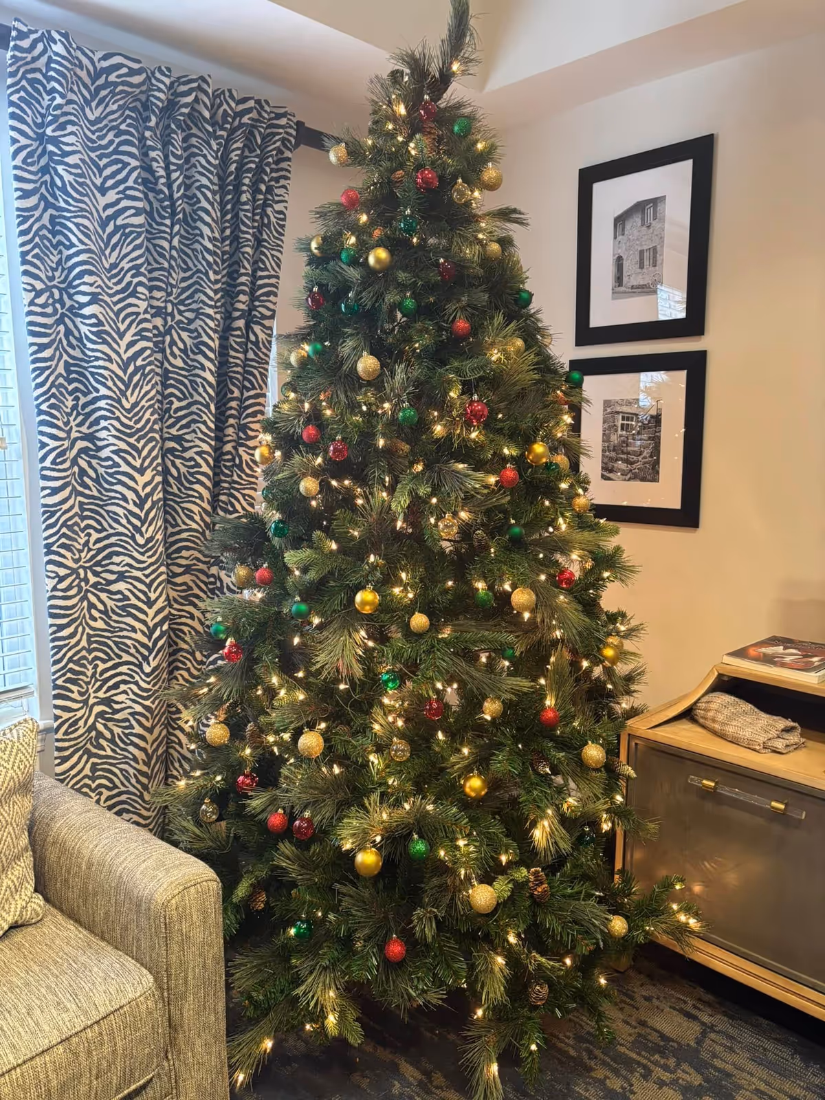 A decorated Christmas tree with red, green, and gold ornaments and white lights stands in a cozy living room corner next to a gray upholstered armchair and a wooden cabinet. Behind the tree, there are two framed black and white photographs on the wall and a window with zebra-patterned curtains.