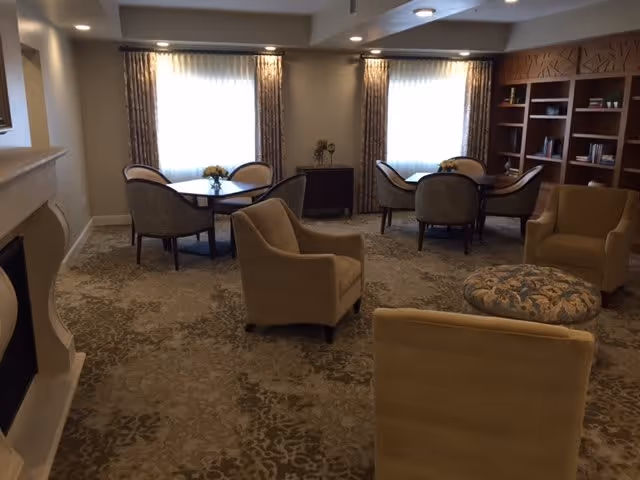 A cozy living room area with beige armchairs arranged around a patterned ottoman. There are two tables with chairs near windows covered by sheer curtains and heavier drapes. A built-in wooden bookshelf is visible on the right side of the room, and a fireplace is on the left.