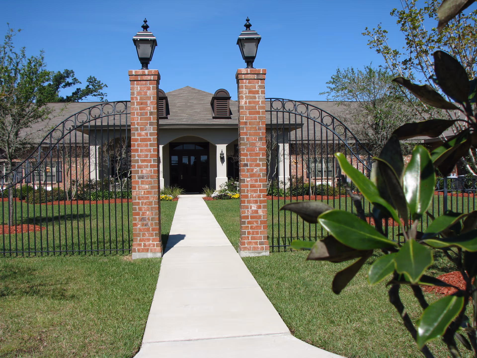 View of the front entrance of a brick building with a concrete walkway leading to double doors. The entrance is framed by two brick pillars topped with lantern-style lights and a black wrought iron gate. Green grass and some trees and shrubs surround the walkway under a clear blue sky.