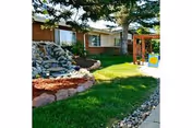 Outdoor garden area at Wheatridge Manor Care Center featuring a green lawn, a rock waterfall feature, trees, and a wooden pergola structure near the building.