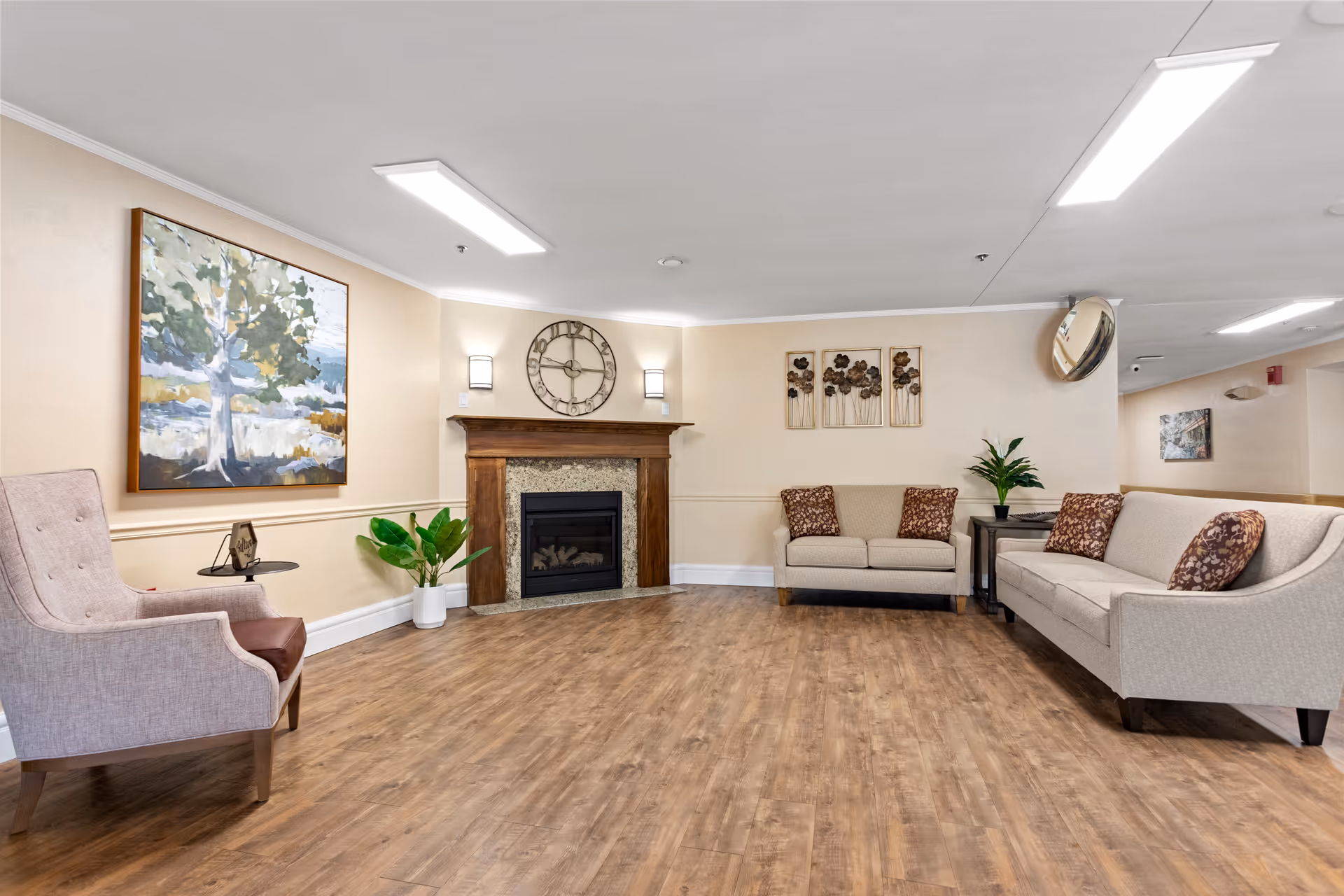A cozy living room area in a nursing care center featuring a wooden floor, a fireplace with a large clock above it, two beige sofas with patterned cushions, a beige armchair, a small side table with a plant, and wall art including a large painting of a tree and a decorative floral piece.