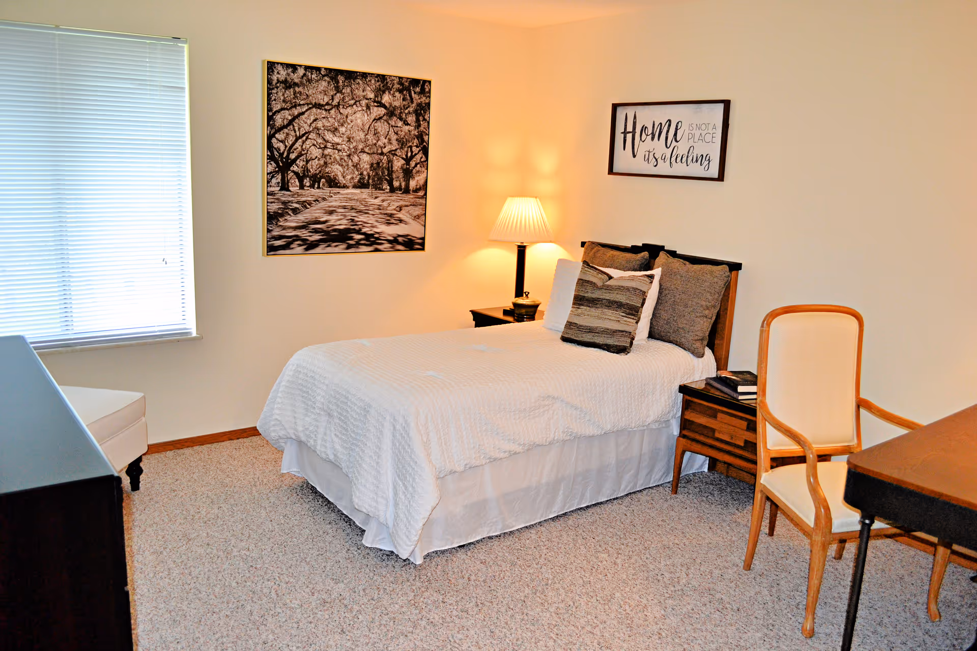 A cozy bedroom with a single bed covered in white bedding and multiple pillows. There is a wooden nightstand with a lamp and books beside the bed. A framed picture of a tree-lined path and a wall sign that reads 'Home is not a place, it's a feeling' hang on the beige walls. A wooden chair with a cushioned seat is placed next to a dark wooden desk. A window with closed blinds lets in natural light.