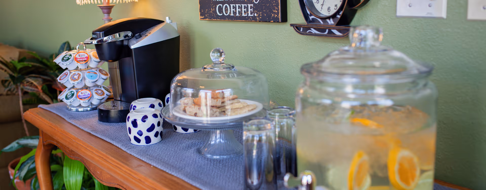 Countertop coffee station with a Keurig, coffee pods, a covered plate of cookies and a glass dispenser of lemon water.