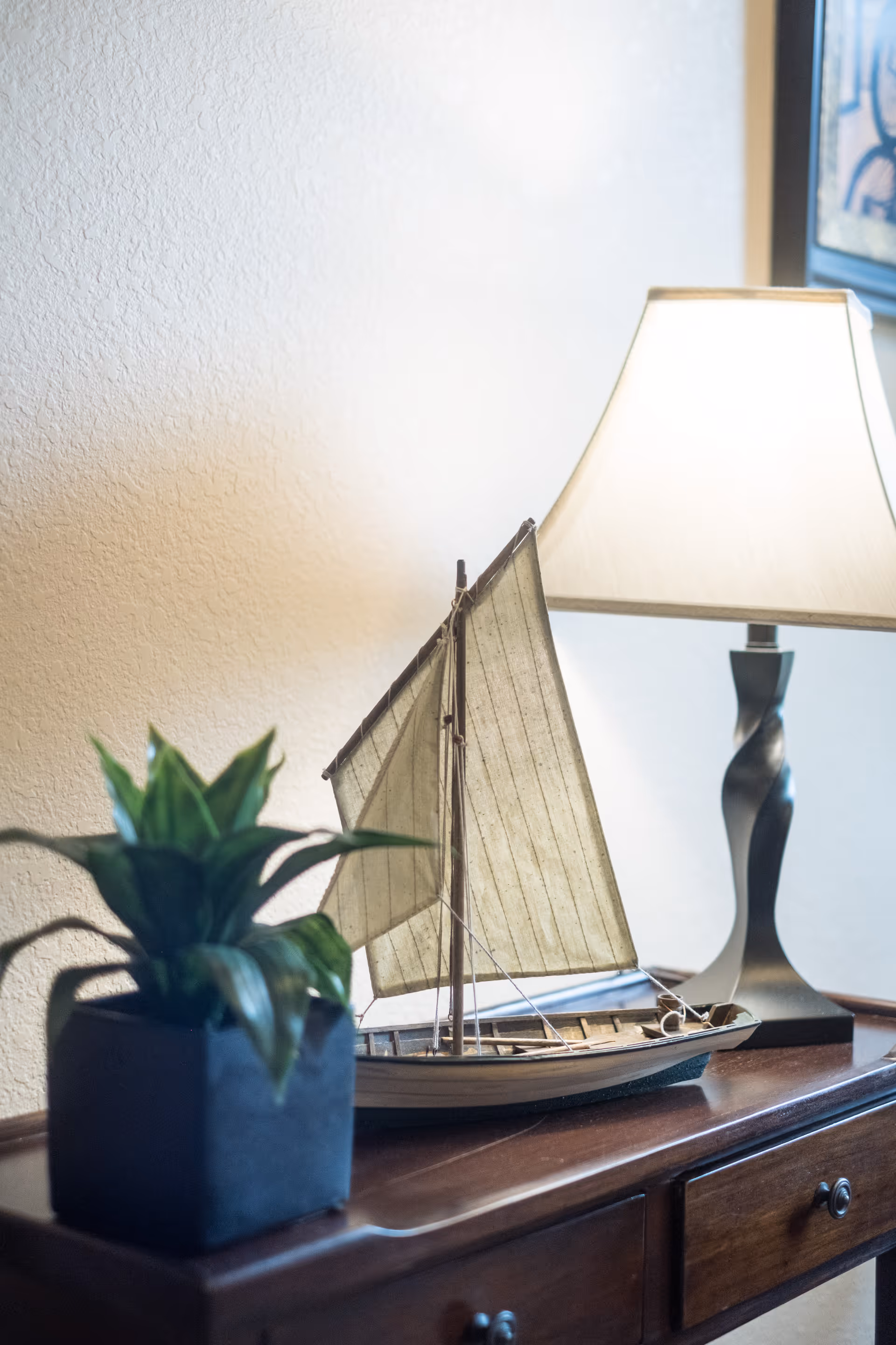 A decorative sailboat model sits on a wooden console table next to a potted plant and a lit table lamp against a textured wall.