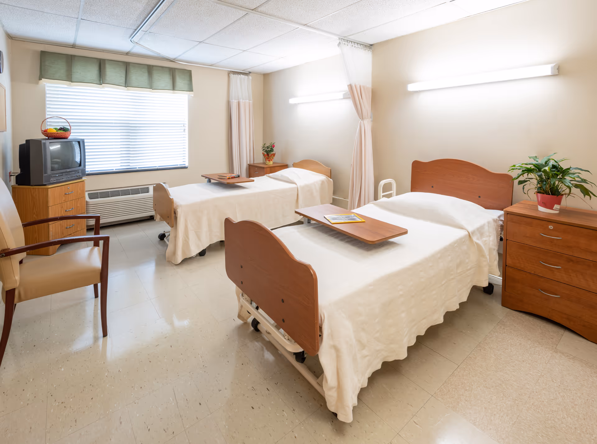 A bright and clean room with two hospital-style beds covered in white bedspreads. Each bed has a wooden overbed table with magazines on top. There is a wooden dresser with a potted plant on it, a small TV on a wooden stand, and a beige armchair. The room has a large window with blinds and a green valance, and the walls are light-colored with fluorescent lighting above each bed.