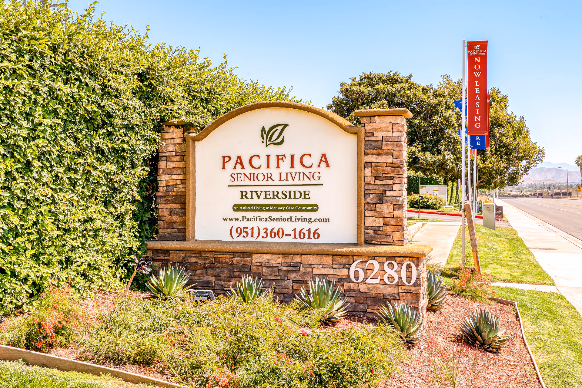 Outdoor stone sign for Pacifica Senior Living Riverside with landscaping and greenery around it, located at 6280. A red vertical banner nearby reads 'Now Leasing'.