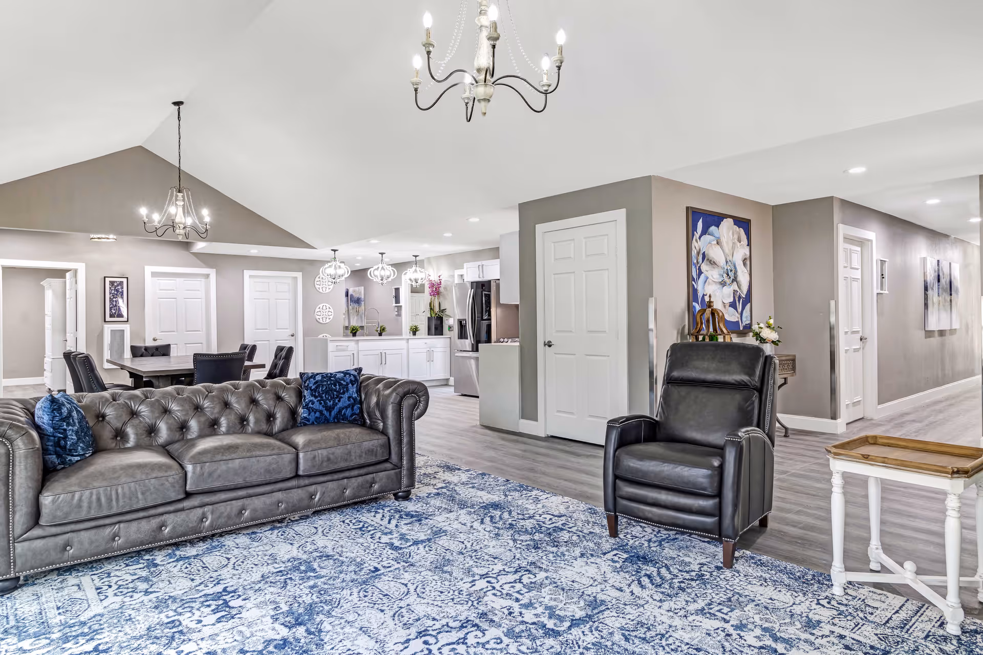 A spacious and well-lit living room area in an assisted living facility featuring a gray tufted leather sofa with blue pillows, a black leather armchair, a white side table with a wooden tray, and a large blue patterned rug. The background shows a dining area with a dark wooden table and chairs, white cabinetry, stainless steel refrigerator, and decorative lighting fixtures. The walls are painted in neutral tones with floral artwork and multiple white doors leading to other rooms.