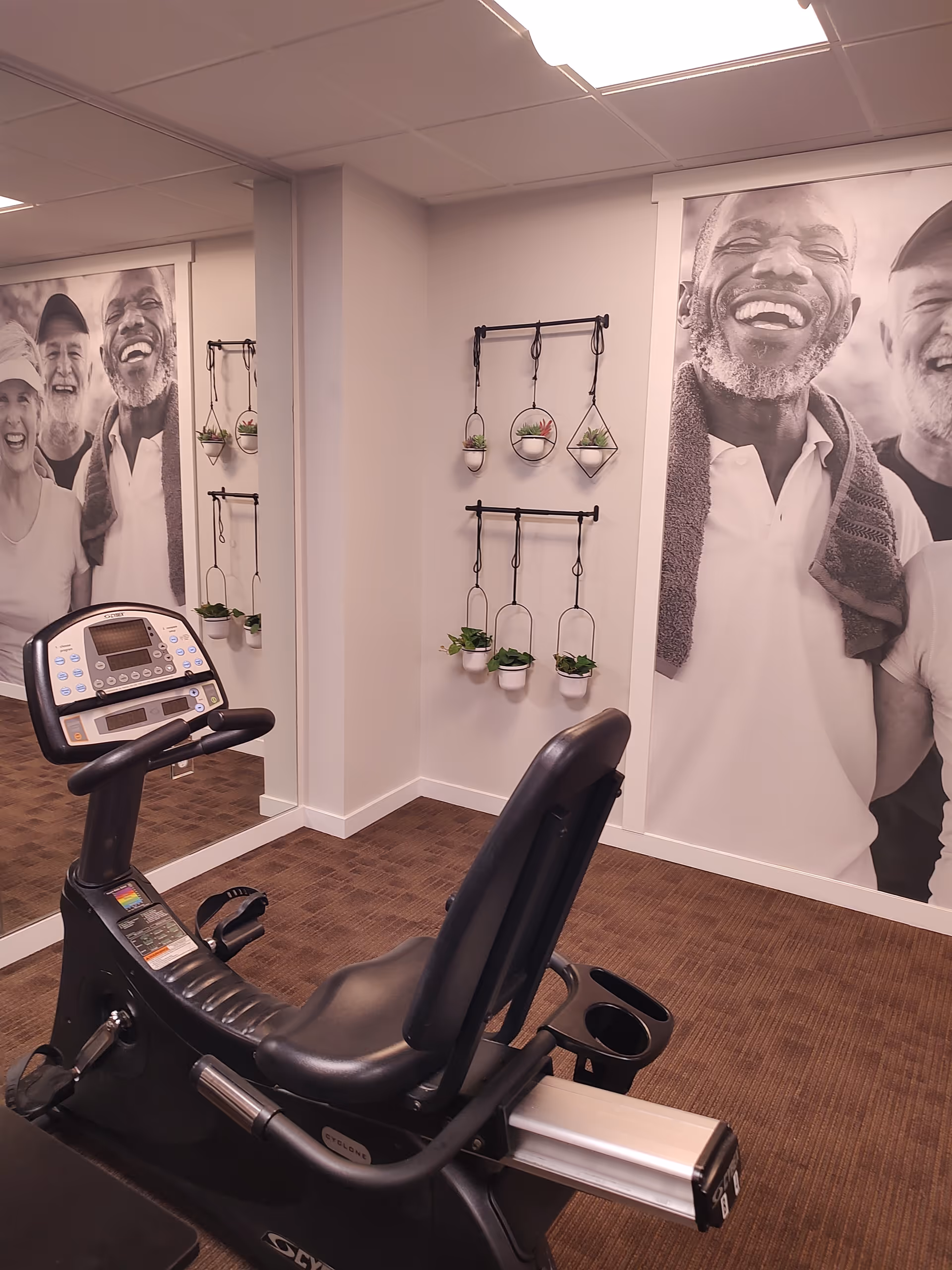 Recumbent exercise bike in a small fitness room with large black-and-white murals of smiling seniors and hanging potted plants.