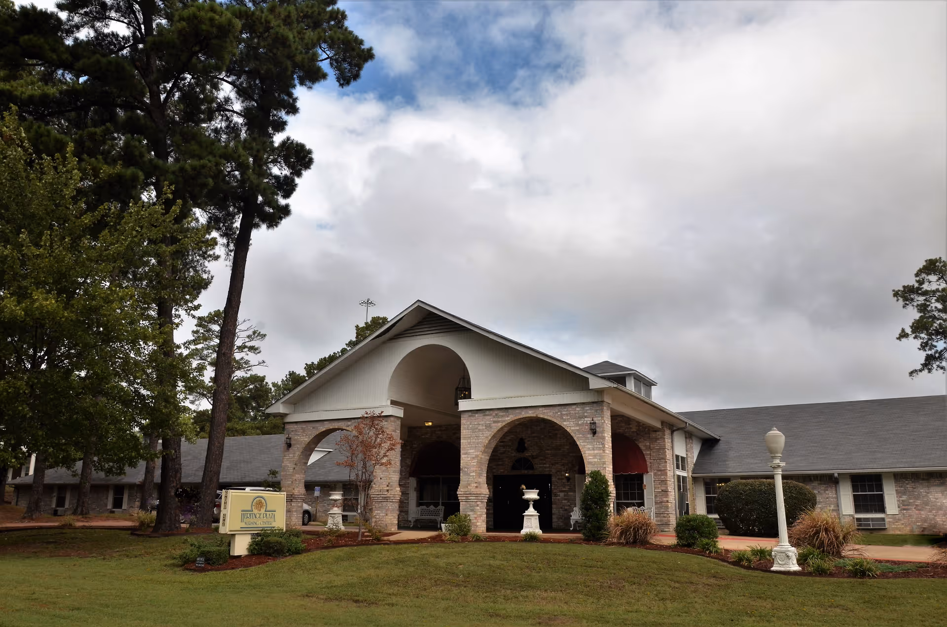 Front entrance of a low brick nursing facility with an arched portico, landscaped lawn, trees and a cloudy sky.