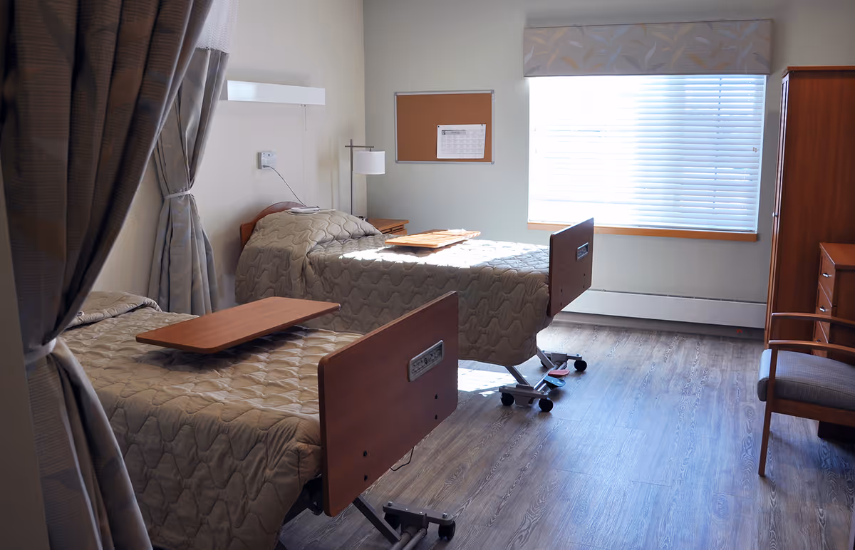 Two hospital-style beds in a bright shared patient room with wood furniture, window blinds, and a curtain divider.