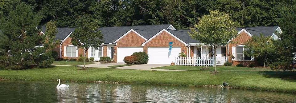 A row of single-story brick residential buildings with white garages and small front porches, surrounded by green lawns and trees, with a pond in the foreground where a white swan is swimming.