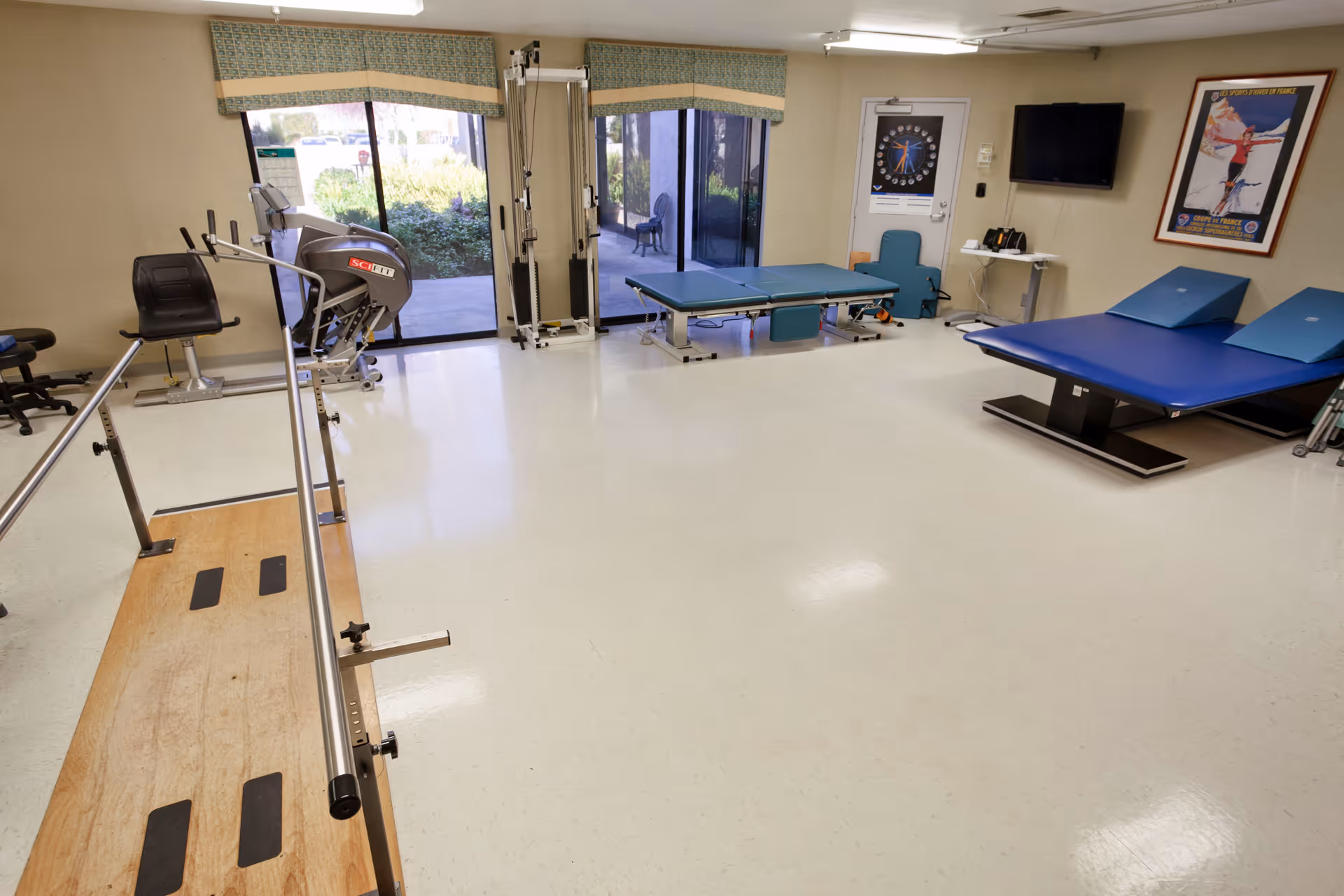Spacious physical therapy room with parallel bars, exercise equipment, treatment tables and sliding glass doors to the outdoors.