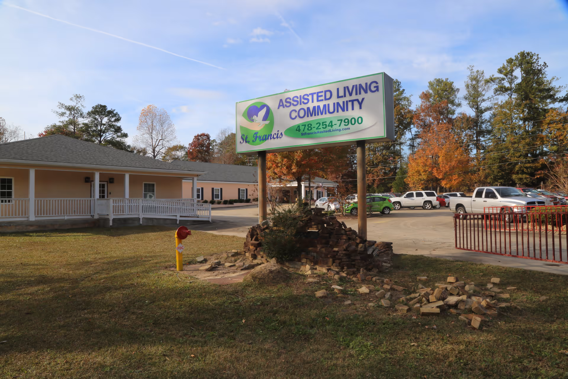Outdoor view of St. Francis Assisted Living Community featuring a large sign with the facility's name, phone number, and website. The sign is mounted on two poles above a small pile of bricks and surrounded by grass. In the background, there are single-story buildings, a parking lot with several vehicles, and trees with autumn foliage under a partly cloudy sky.