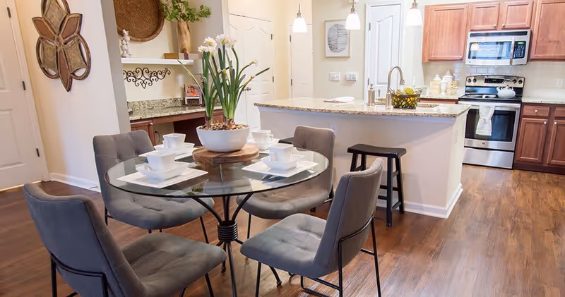 A modern kitchen and dining area with a round glass table set with white dishes and four gray cushioned chairs. The kitchen features wooden cabinets, stainless steel appliances including a microwave and oven, a granite countertop island with a fruit basket, and pendant lights hanging above. The floor is wooden, and decorative items are placed on shelves and walls.