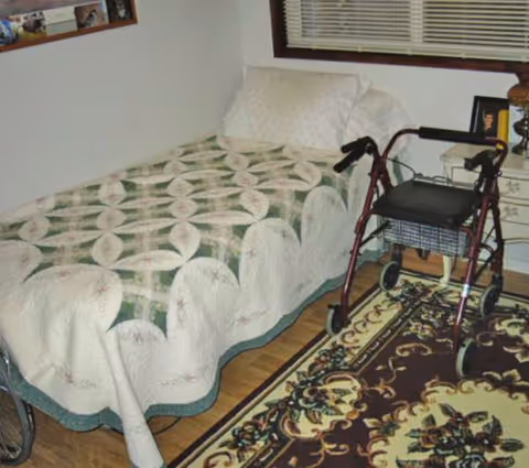 Bedroom in a senior facility featuring a quilted bed and a walker.