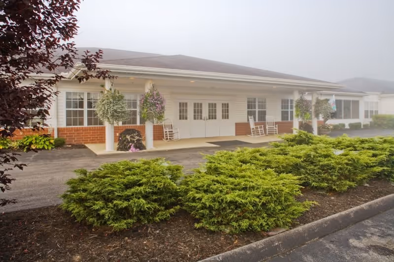Front exterior view of a single-story building with white siding and brick accents. The entrance features white double doors flanked by large windows and white columns with hanging flower baskets. There are white rocking chairs on the porch, and green shrubs line the driveway in front of the building. The sky is overcast or foggy.