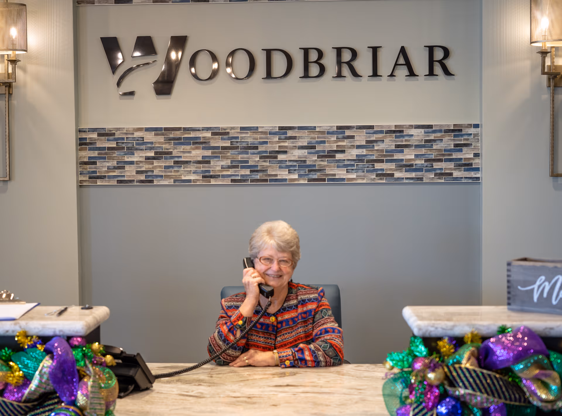 An elderly woman with short gray hair and glasses is sitting behind a reception desk, smiling and talking on a telephone. Behind her is a wall with the word 'WOODBRIAR' displayed in large letters above a decorative tile strip. The reception desk is decorated with colorful Mardi Gras-themed ribbons and ornaments.