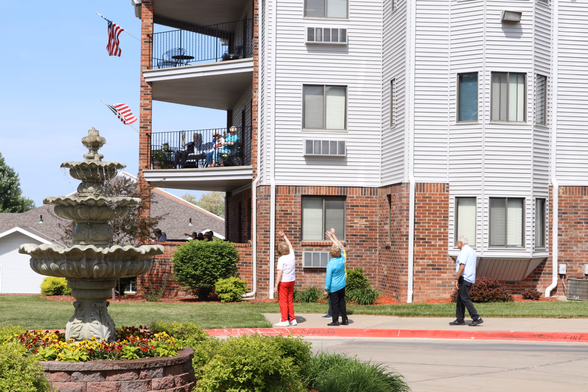 Front of a senior living building with a decorative fountain in the foreground and residents walking and sitting on balconies.