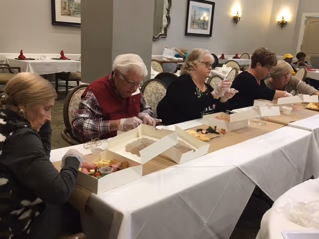 A group of elderly people sitting at a long table in a dining area, each with a box of food in front of them. They appear to be engaged in an activity involving the food, wearing gloves and focused on their tasks. The room has framed pictures on the walls and tables set with white tablecloths and red napkins in the background.
