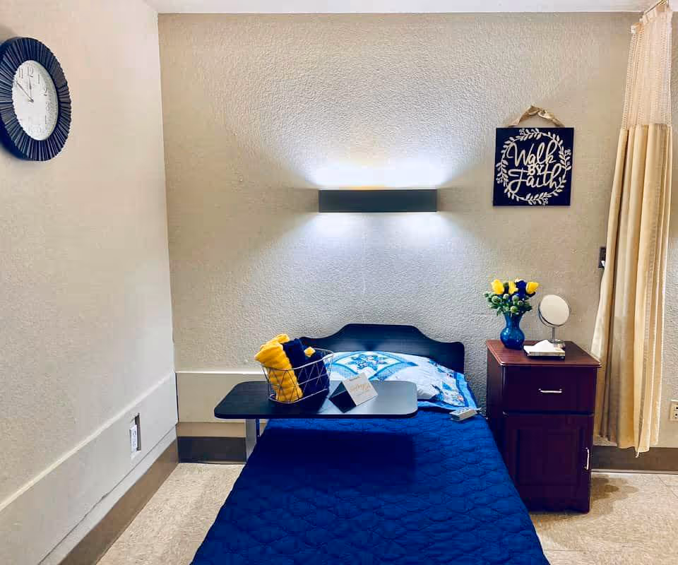 A single bed with a blue quilt and a decorative pillow is placed against a beige wall. Above the bed is a modern wall light and a framed sign that reads 'Walk by Faith.' A small wooden nightstand with a vase of yellow and blue flowers, a small mirror, and tissues is next to the bed. A black overbed table holds a basket with yellow and blue towels and a small card. A round wall clock is mounted on the adjacent wall, and beige curtains hang to the right.
