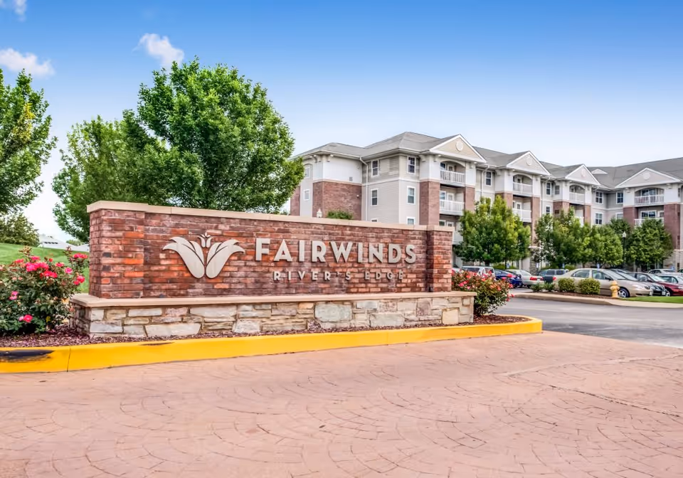 Entrance sign for Fairwinds River's Edge senior living facility made of brick and stone with a flower logo, surrounded by landscaping with trees and flowers, and a multi-story building with balconies and parked cars in the background under a clear blue sky.