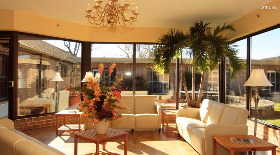 A bright and cozy atrium with large floor-to-ceiling windows letting in natural light. The room features beige sofas arranged around wooden tables, a large potted palm plant, table lamps, a chandelier, and a floral arrangement on a central table. Outside the windows, a brick building and outdoor seating area are visible.