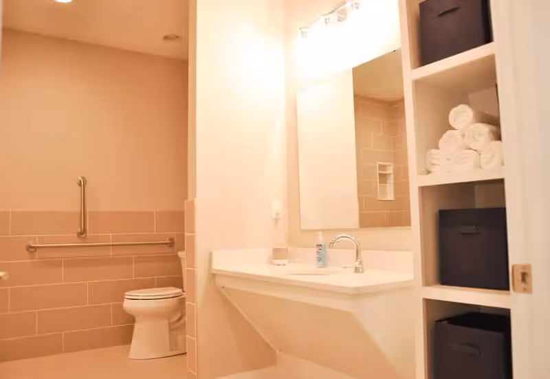 Accessible bathroom with a wall-mounted sink and mirror, toilet with grab bars, and shelving holding towels and storage bins.